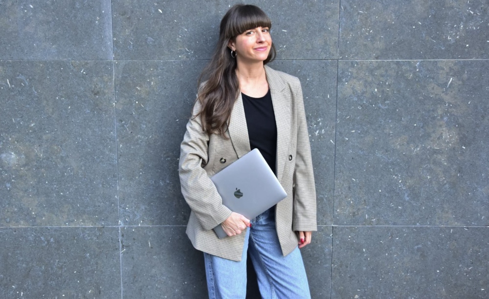 A woman with brown hair posing for a business photo, holding a Macbook, wearing a light brown jacket and jeans